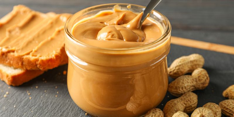 Jar of creamy peanut butter surrounded by peanuts and bread with peanut butter spread.