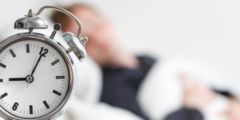 an alarm clock with someone sleeping in the background